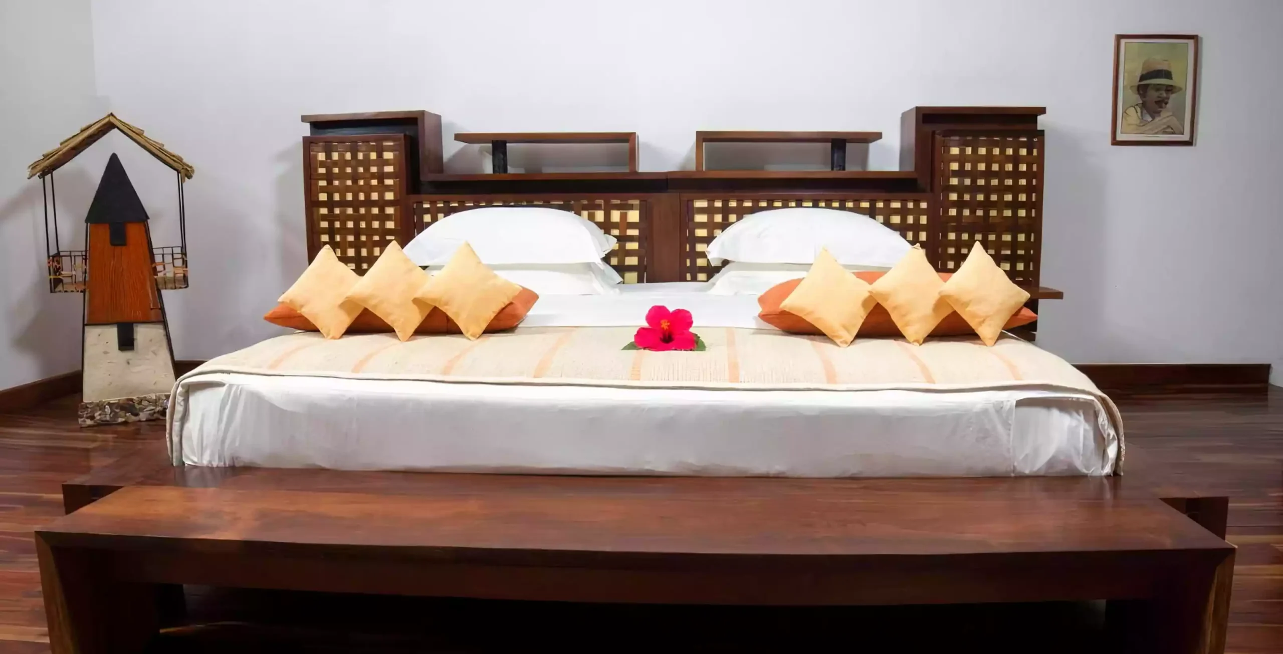 Large bed with wooden headboard, six orange and yellow cushions, beige throw, and a red flower on top.