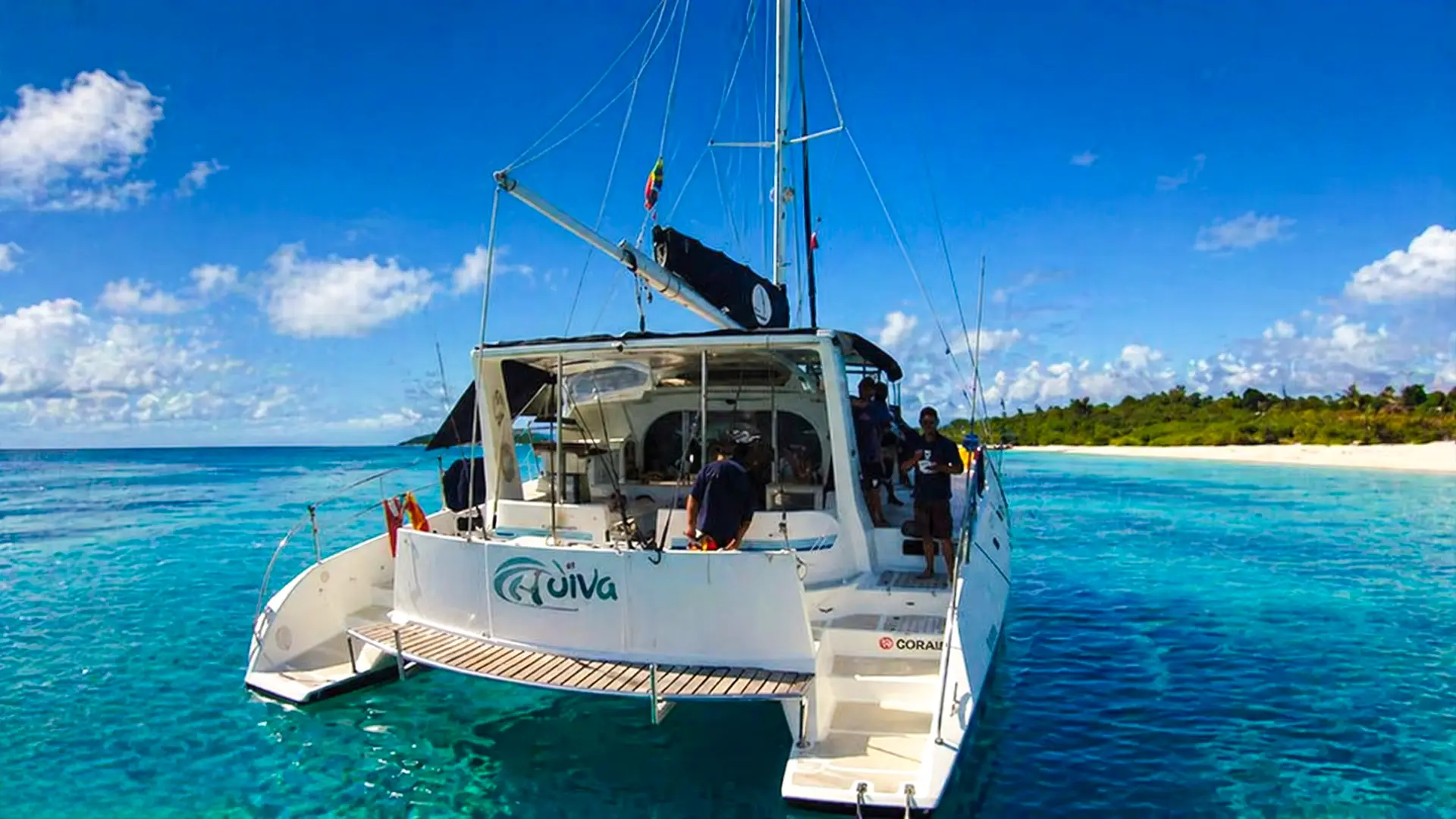White sailing catamaran named Adiva anchored in clear blue water near a tropical beach with people onboard.