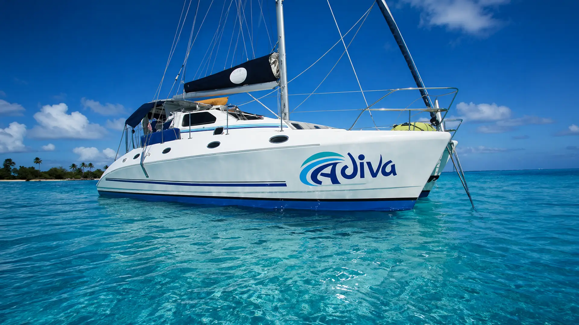 White sailboat named Adiva floating on clear turquoise water under a bright blue sky.