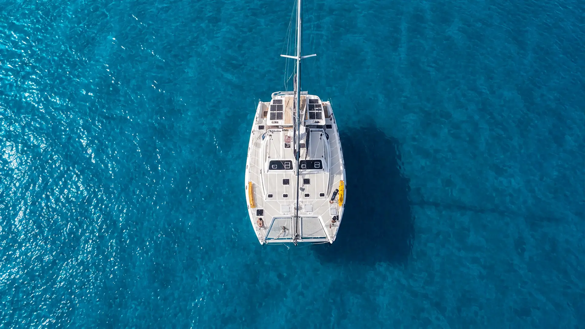 A white catamaran sailboat floats on clear turquoise blue water seen from directly above.
