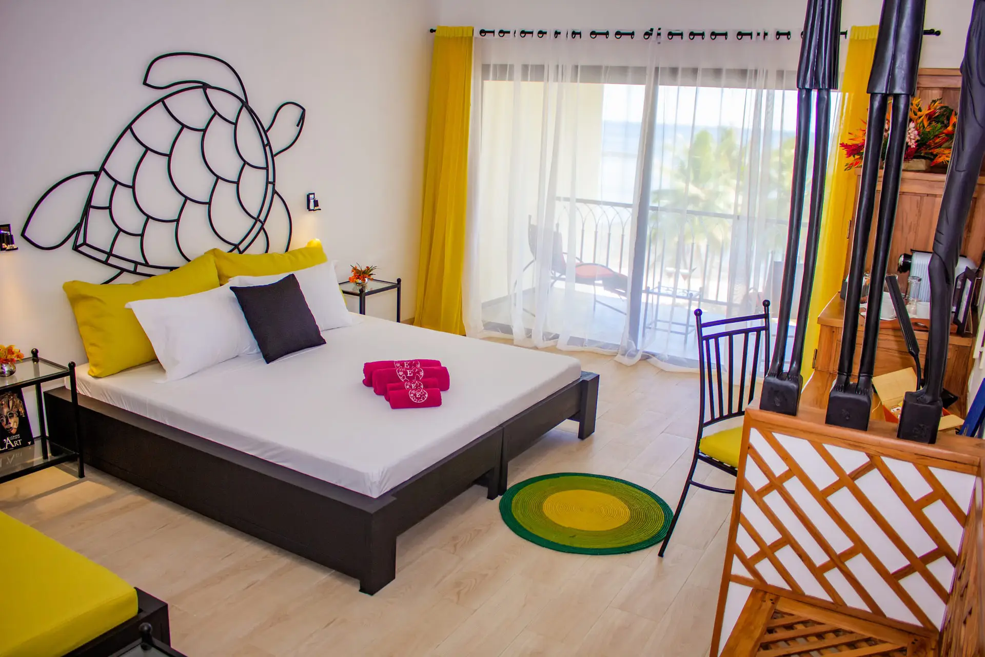 Bright hotel room with a bed, yellow pillows, towel set, turtle wall art, and balcony with sheer curtains.