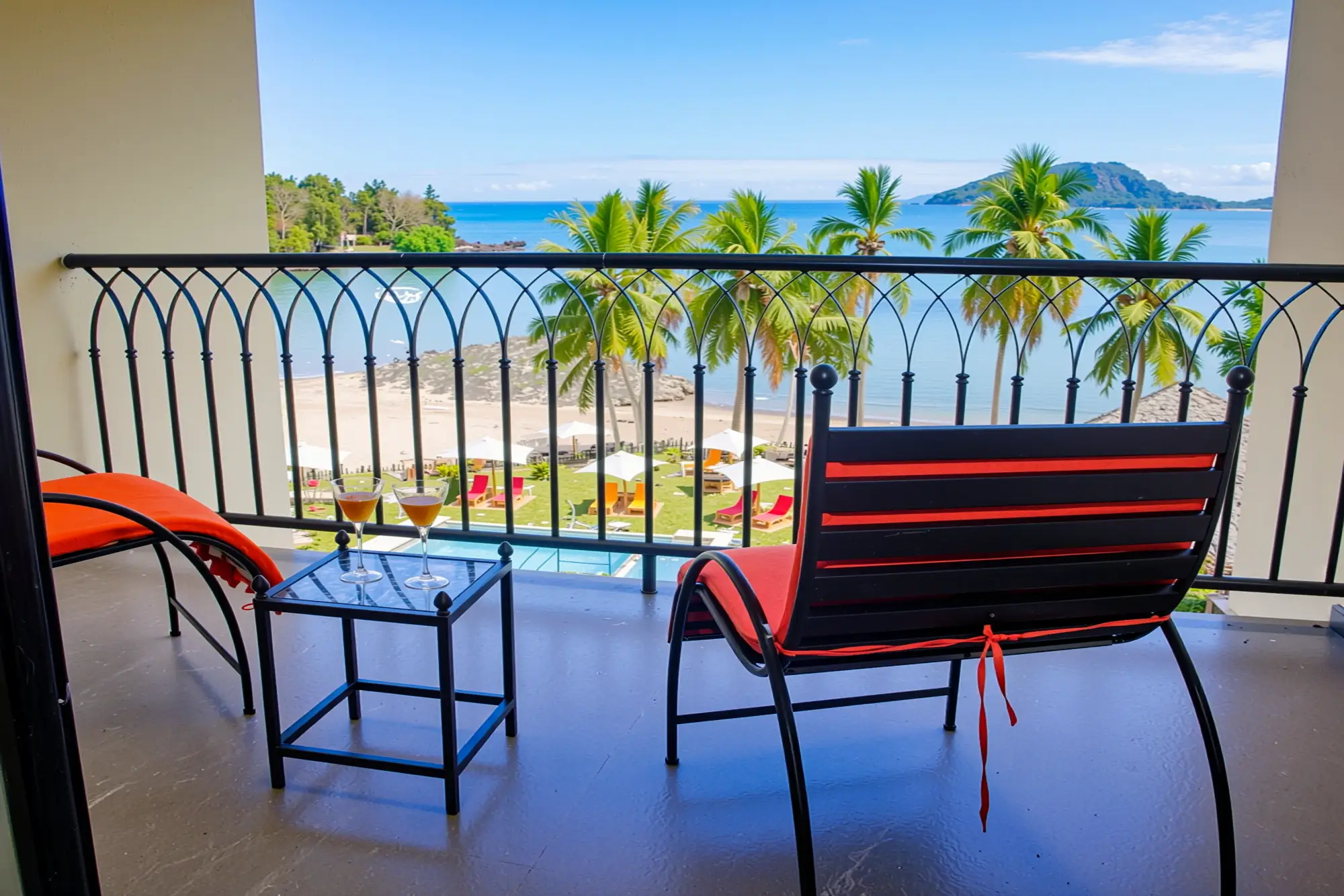 Balcony view with two lounge chairs and drinks overlooking palm trees, beach, ocean, and island.