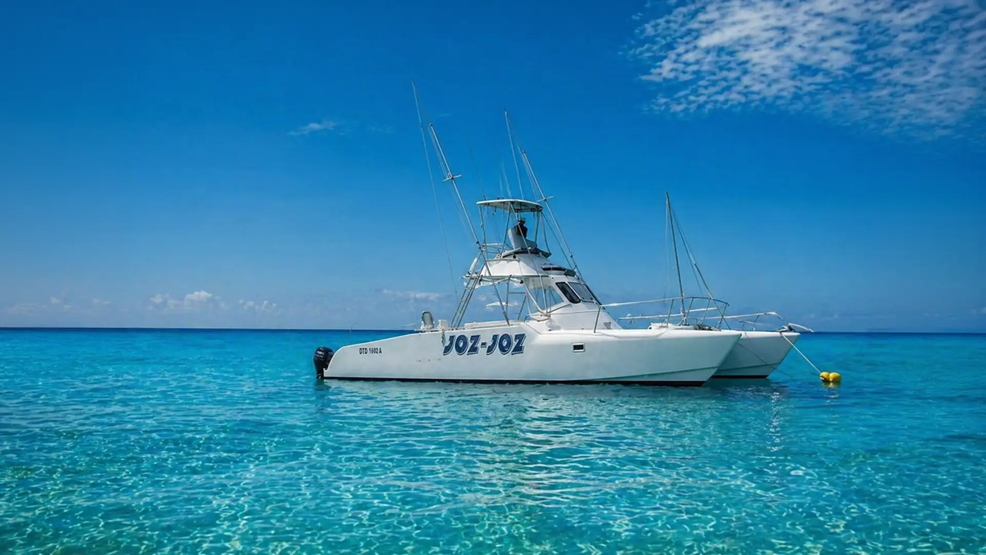 White fishing catamaran named JOZ-JOZ floating on clear blue ocean water under a partly cloudy sky.
