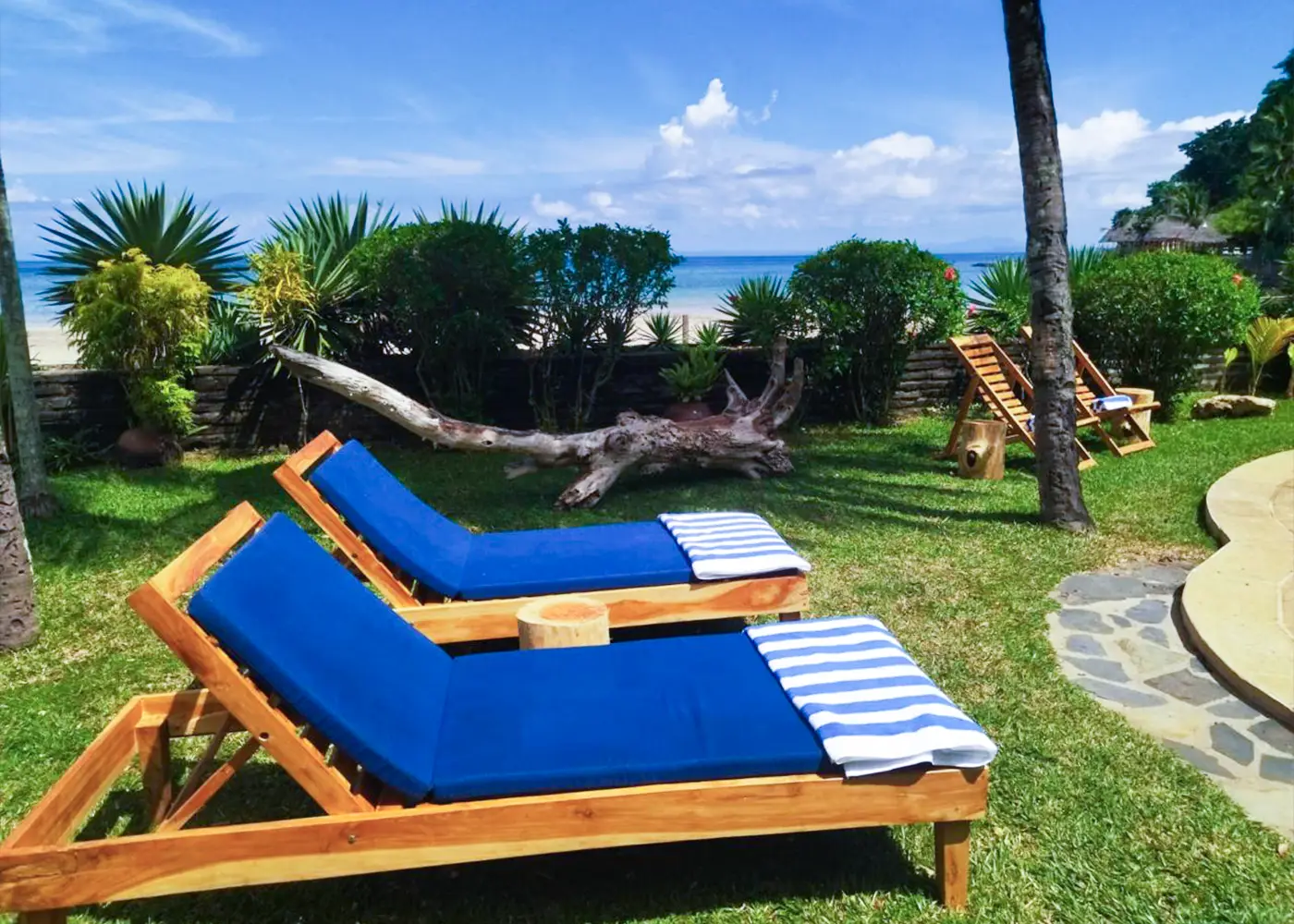 Two blue lounge chairs with striped towels on green grass by a tropical beach under clear sky.