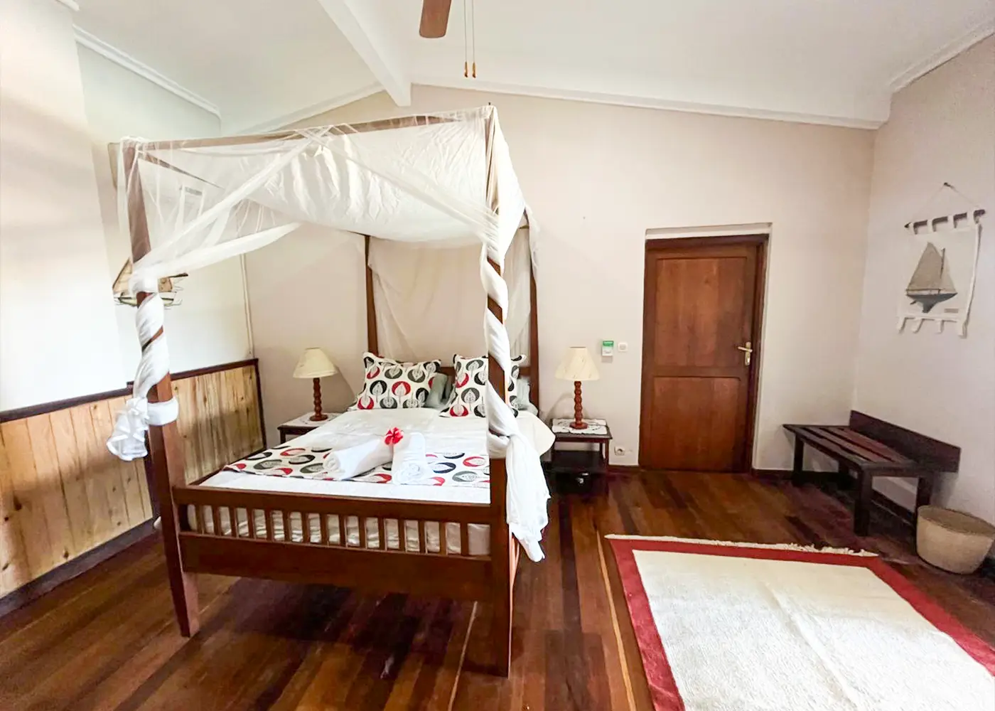 Wooden four-poster bed with white drapes and patterned pillows in a room with wooden floor and door.