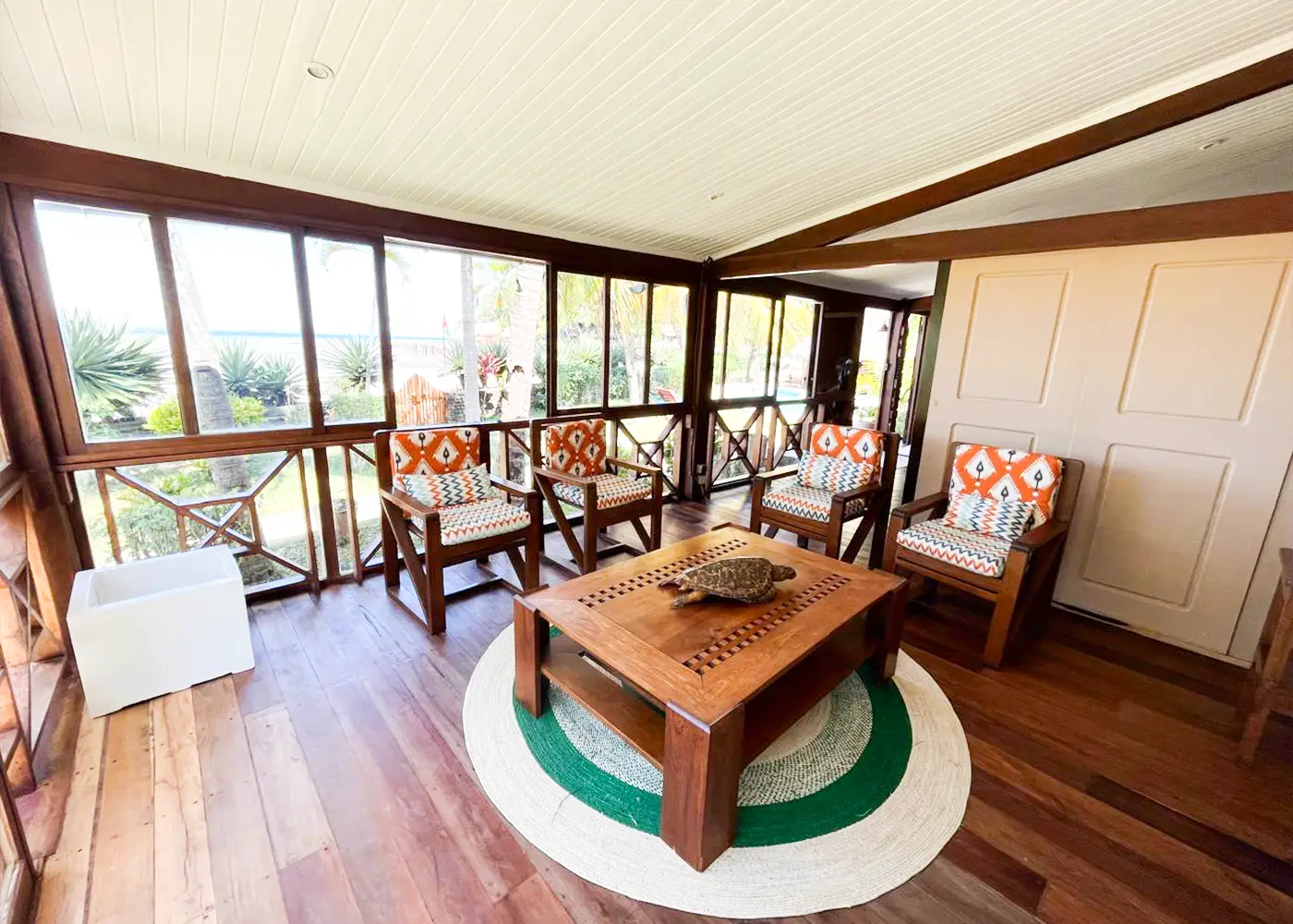 Sunroom with wooden floor, glass windows, four wooden chairs with orange patterned cushions, and a wooden table.