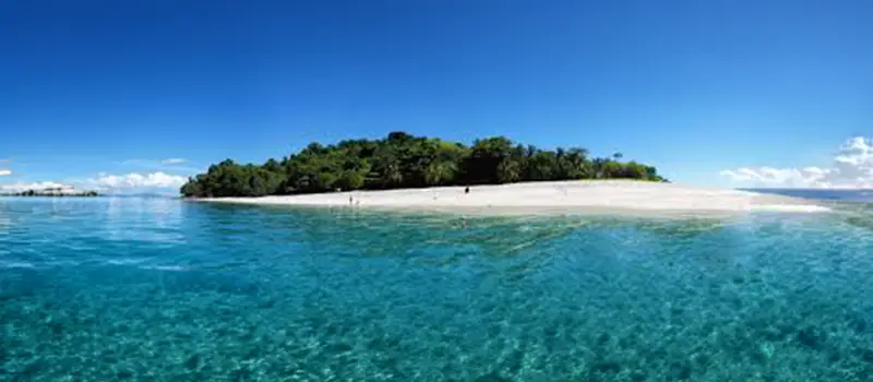 Small island with dense trees and white sandy beach surrounded by clear turquoise ocean under blue sky.