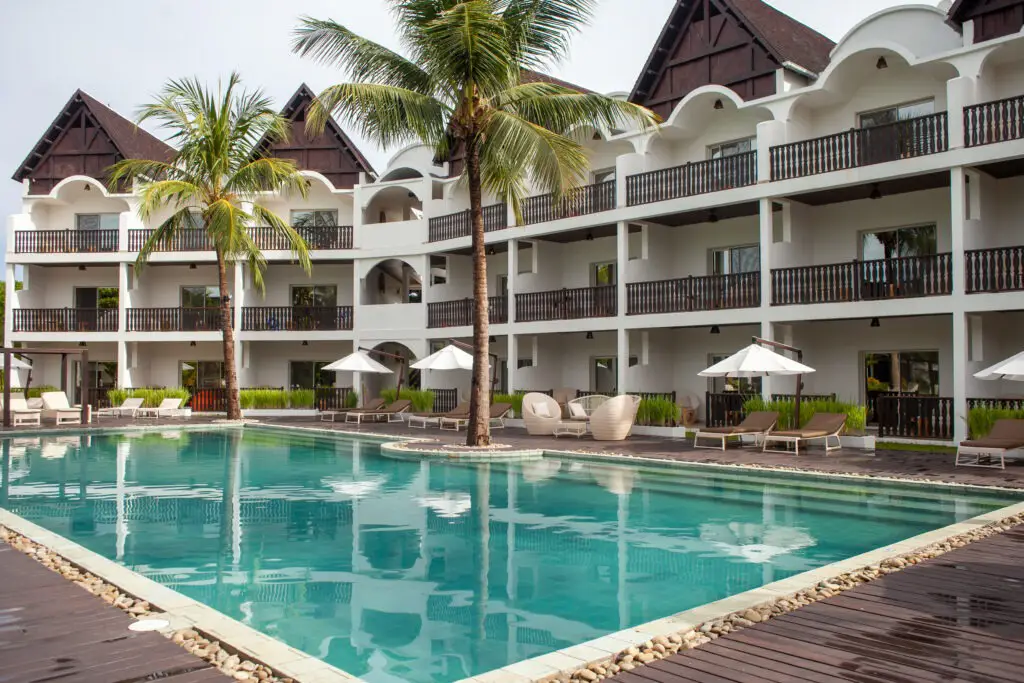 Resort-style swimming pool with lounge chairs, umbrellas, palm trees, and a white three-story building.