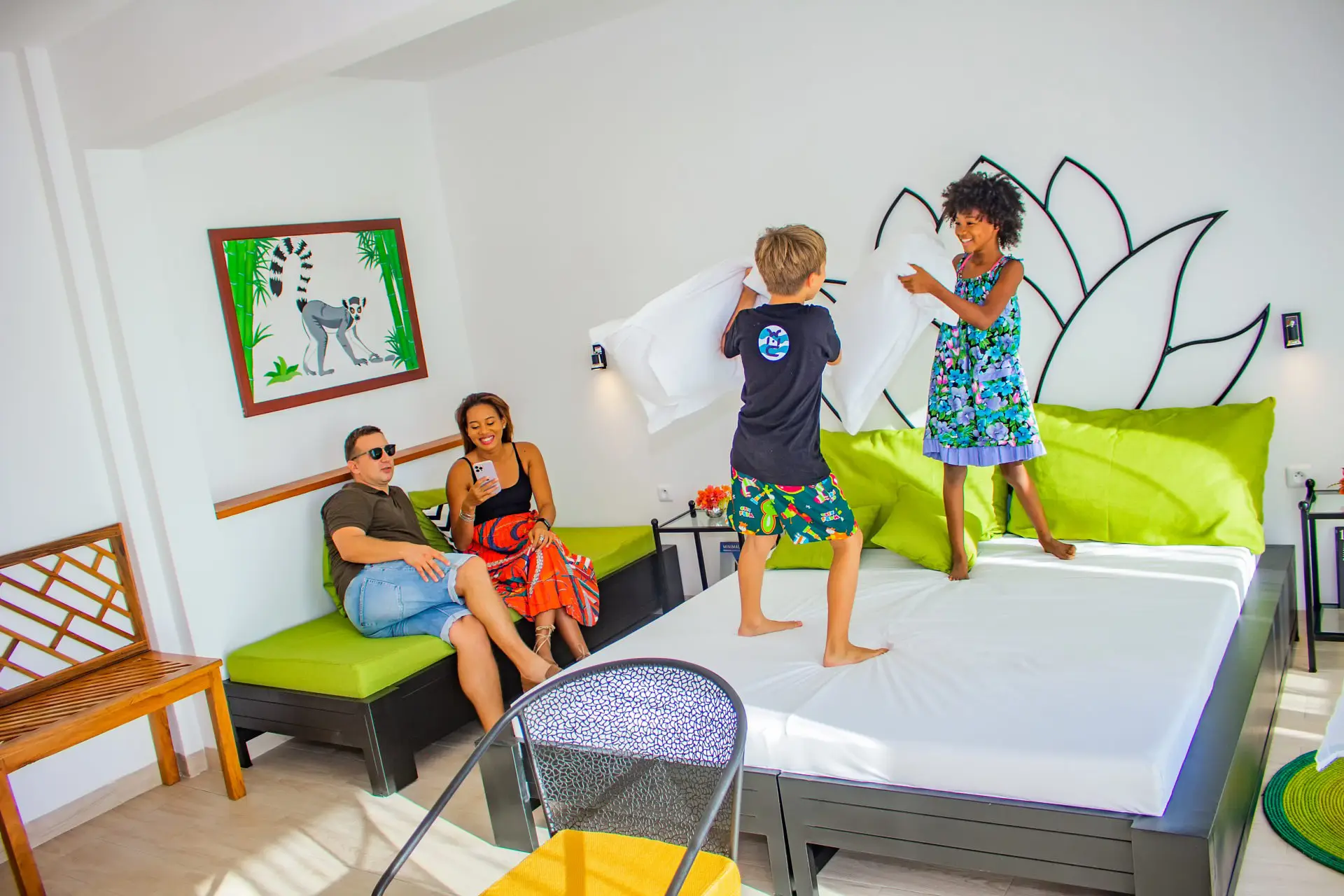 Two kids having a pillow fight on a bed while a couple sits on a green bench smiling and looking at a phone.