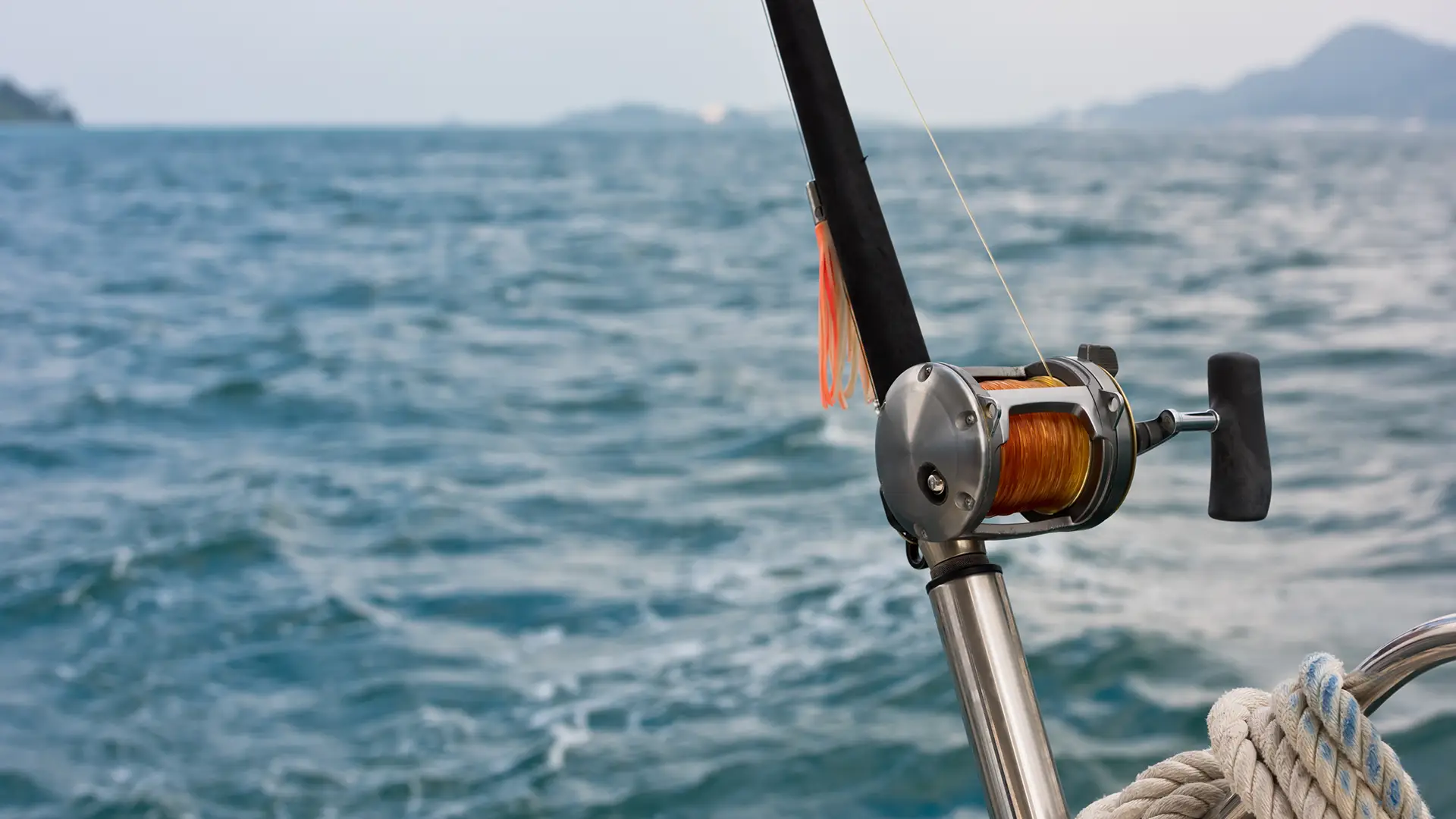 Close-up of a fishing rod and reel mounted on a boat above blue ocean water with distant land on horizon.