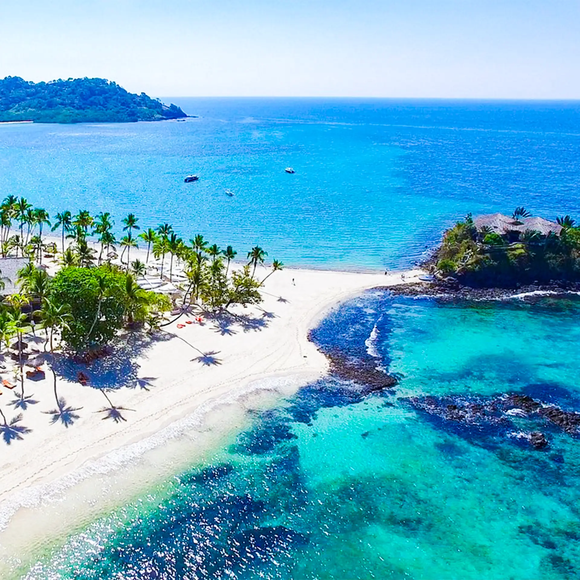 Aerial view of a tropical island beach with palm trees, white sand, clear turquoise water, and boats offshore.