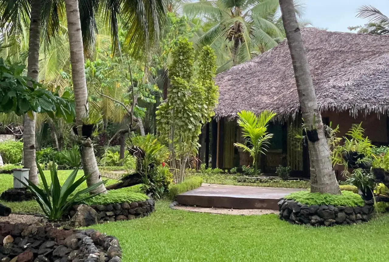 Thatched roof hut surrounded by lush tropical plants, palm trees, and manicured green grass.
